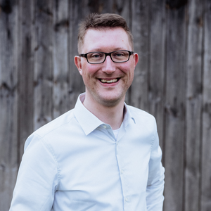 Smiling person wearing glasses and a light blue button-down shirt, standing in front of a wooden fence.
