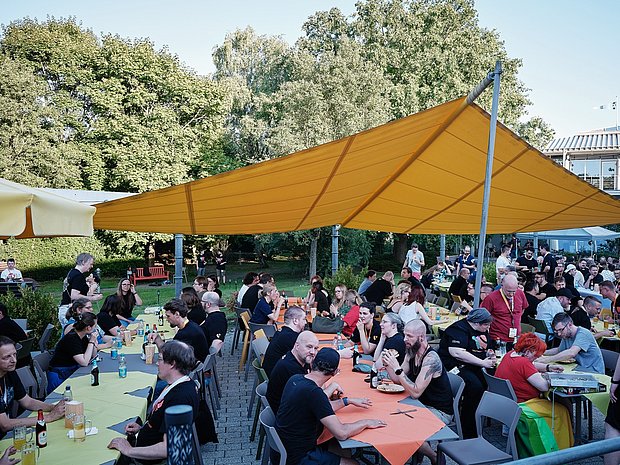 Groups of people sitting at outdoor tables under a large yellow canopy during the social night at TYPO3 Developer Days, chatting, eating, and enjoying drinks in a garden setting.