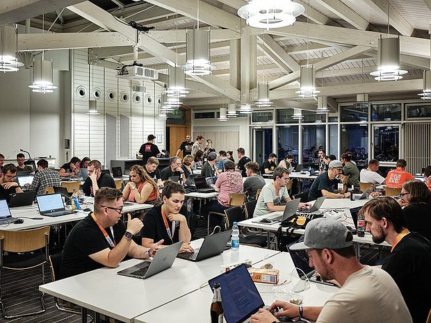 Large group of developers working on laptops at long tables during the coding night at TYPO3 Developer Days in a bright, open hall.