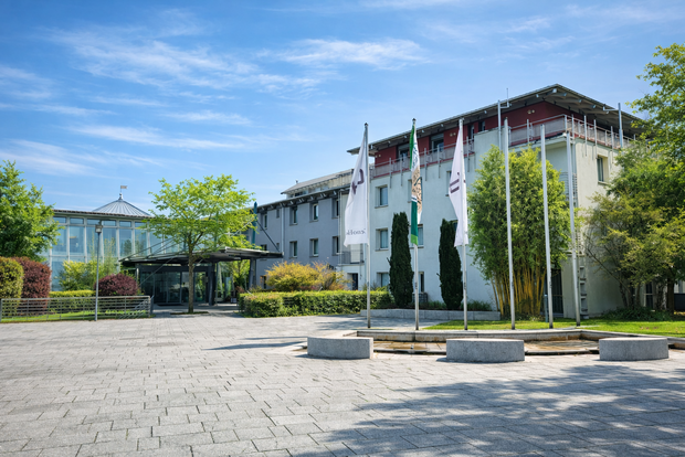 Exterior view of the Geno Hotel in Karlsruhe on a sunny day, with flagpoles in front, modern buildings in the background, and trees and greenery around the entrance.