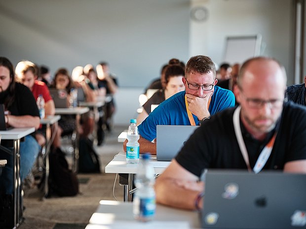 Rows of developers seated at desks, focused on their laptops during the TYPO3 Developer Days certifications, with water bottles on the tables and sunlight coming through the room.