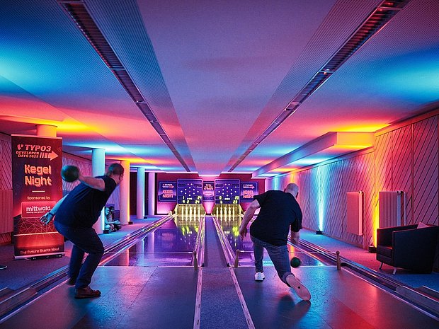Two people bowling on adjacent lanes in a neon-lit bowling alley during the TYPO3 Developer Days Bowling Night, with colorful purple and blue lights reflecting on the floor.