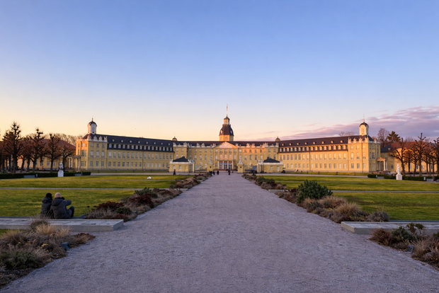 Karlsruhe Palace seen from the central garden path at sunset, with the symmetrical yellow facade and towers under a clear sky.