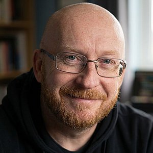 Smiling bald person with glasses and a short beard, sitting at a desk in a home office with computer monitors and a bookshelf in the background.